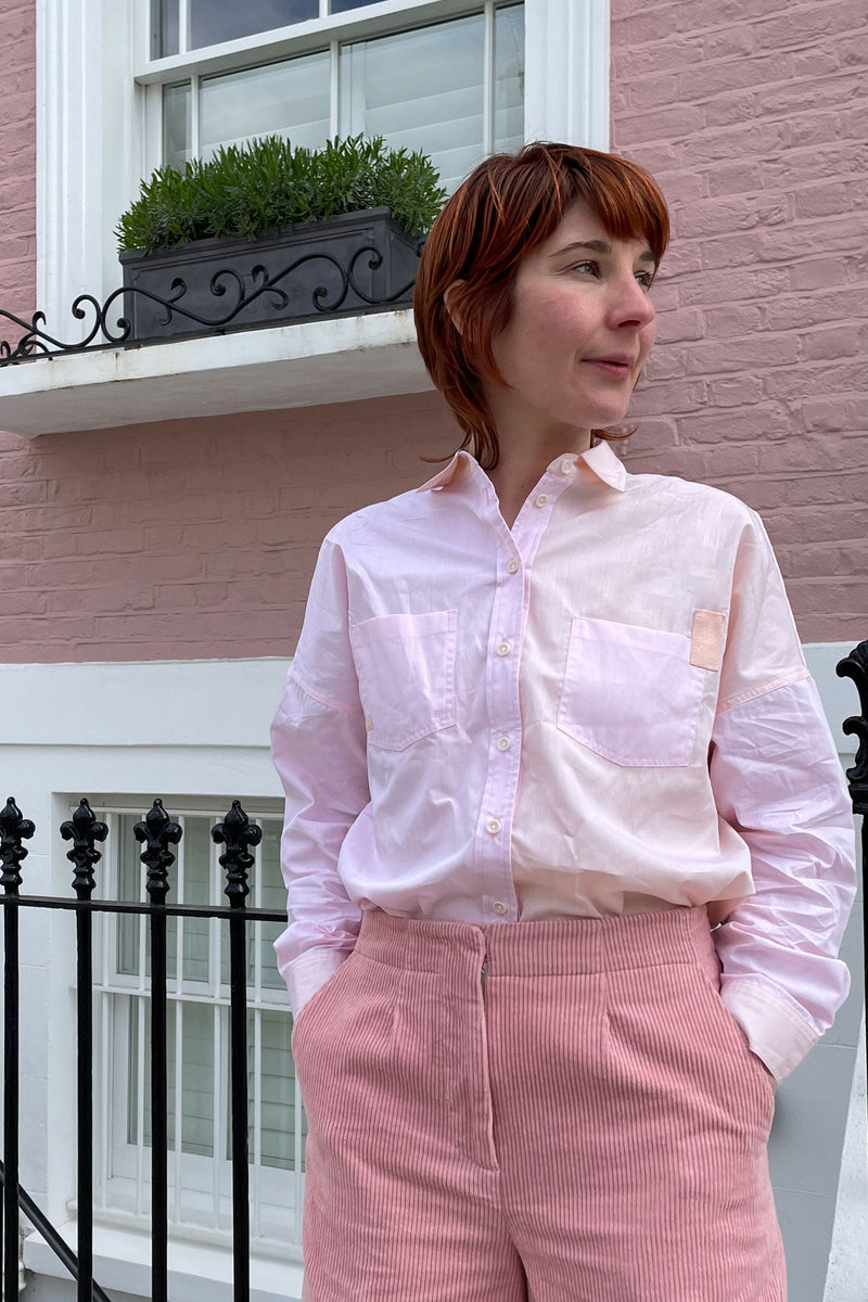 Woman stands in front of a pink brick house, wearing Saywood's boxy silhouette Lela Shirt in pastel pink and orange, tucked into a pair of pink corduroy trousers.