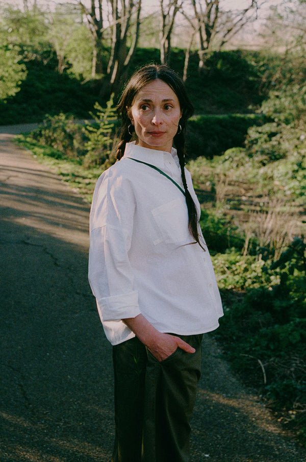 A woman is standing on a path in a green park surrounded by nature. She is turned to the side looking over her shoulder with her hand in her trouser pocket. She wears Saywood's white recycled cotton Lela Boxy Shirt with the khaki Amelia Wide Leg Trousers. Her hair is plaited in pigtails and she has a green cross body bag across her.