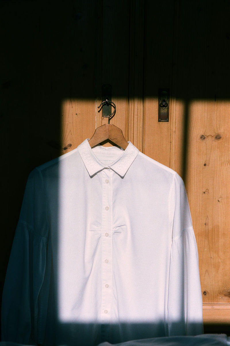 Saywood's Edi Volume Sleeve shirt with lace trim collar is hanging on a wooden hanger on an old French pine wardrobe. The room bathed in sunlight a square of direct sunlight from the window opposite can be seen shining on the shirt, with the surround in darker shade. The soft gathers at the bust on the shirt can be seen as a detail.