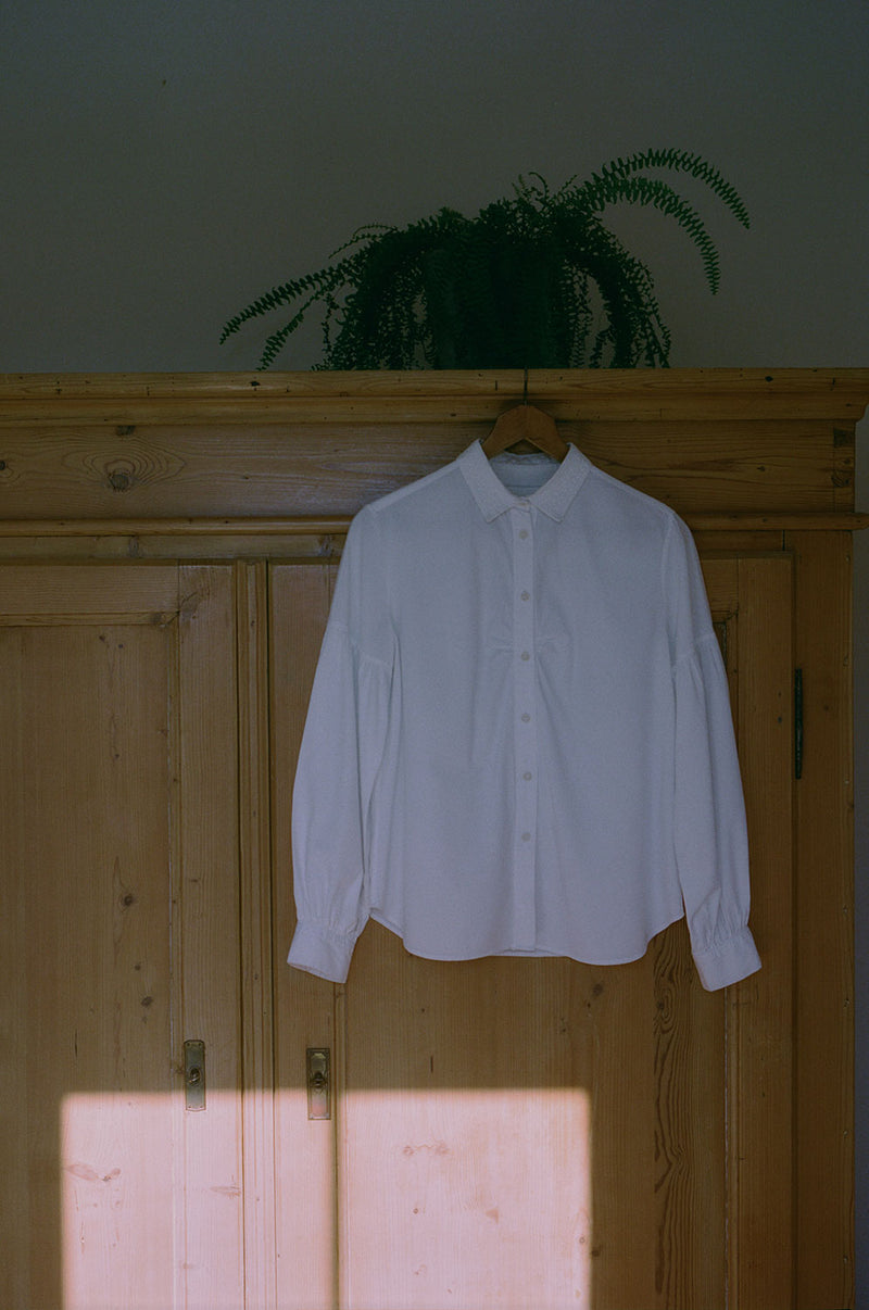 Saywood's Edi Volume Sleeve shirt with lace trim collar is hanging on a wooden hanger on an old French pine wardrobe. There is a fern plant on top of the wardrobe. The image is mostly in shade, with the room bathed in sunlight a square of direct sunlight from the window opposite can be seen at the bottom of the image.