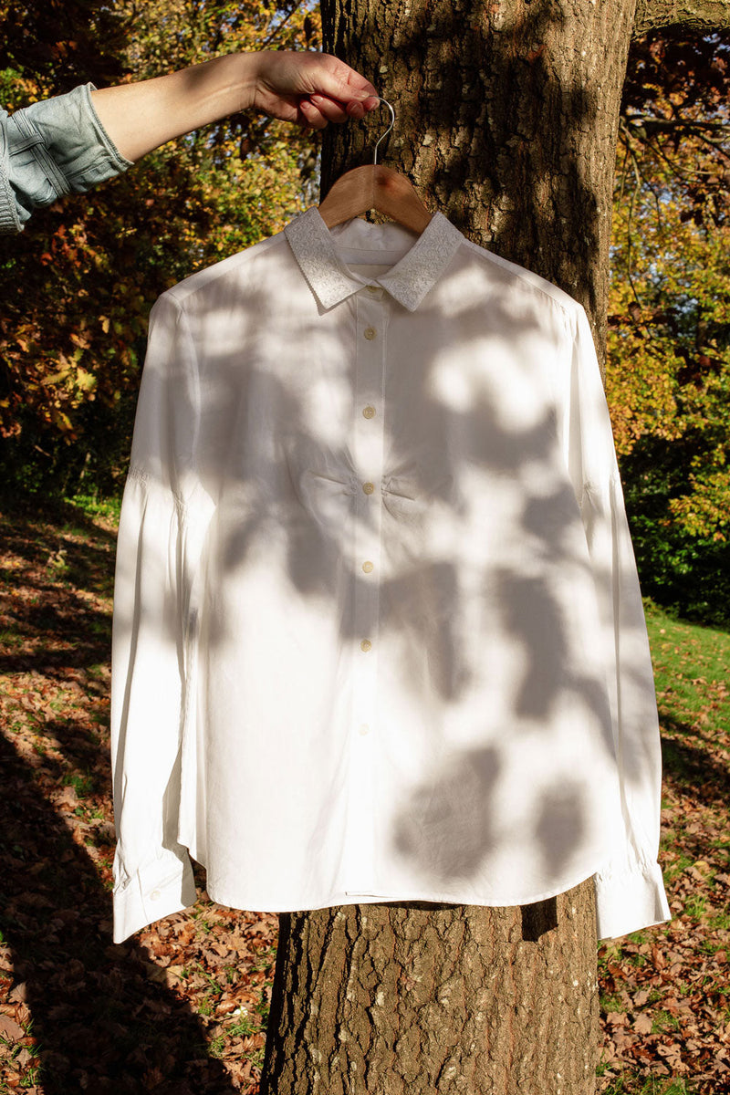 Women's white shirt with lace collar, made in England, on a wooden hanger, being held by a hand in front of a tree. Shadows f leaves can be seen across the shirt from the light.