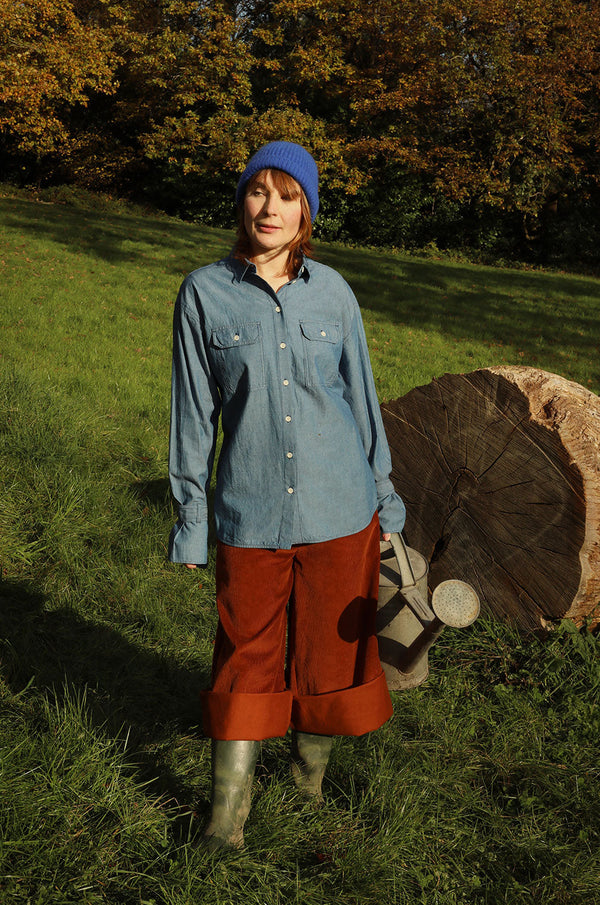 A woman standing in a green field with trees behind her and a big tree trunk in the background. She is wearing Saywood's Caramel Tan Corduroy Trousers in Organic Cotton, styled rolled up with wellies, with the cotton Zadie Boyfriend Shirt in Japanese Denim, and a blue beanie hat. She holds a watering can.