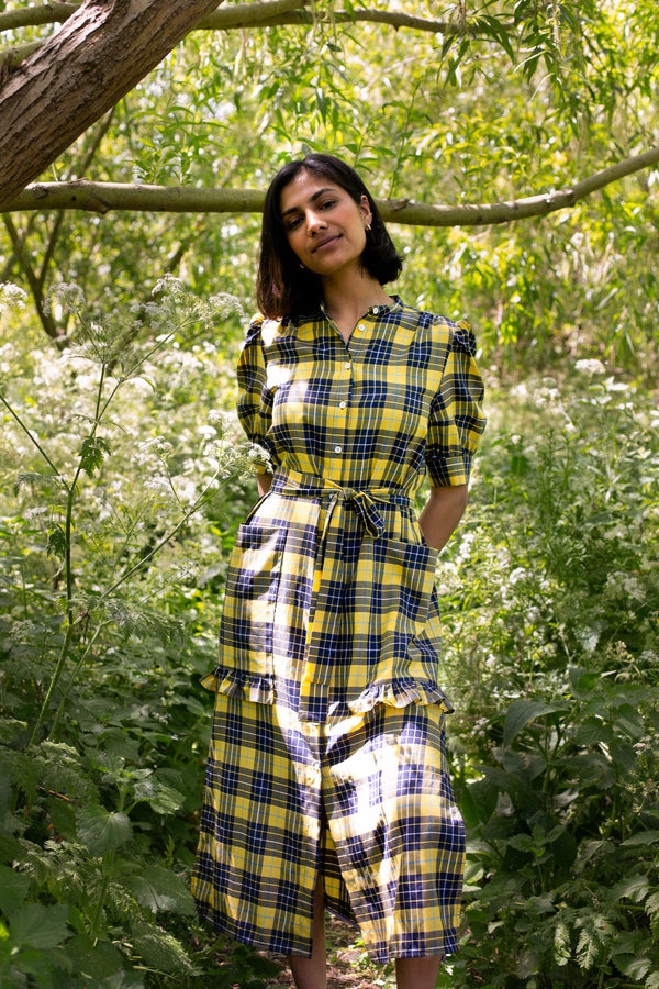 Model stands by a tree surrounded by greenery, wearing Saywood's Rosa yellow check shirtdress, with belt loosely tied round the waist. She smiles and her hands are behind her back, and puff sleeves and patch pockets with ruffles are seen on the dress.