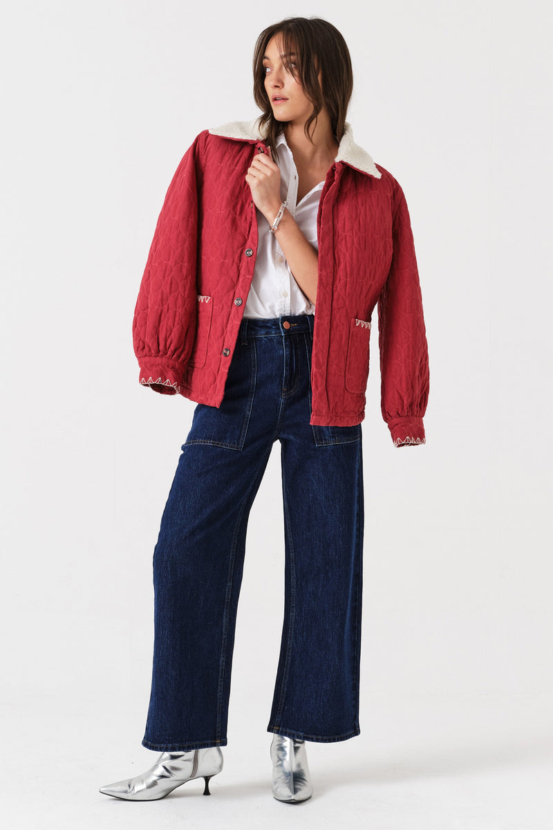 Woman wearing dark wash blue cropped wide-leg jeans with pocket details, a white shirt, and a quilted red jacket with a sherpa collar on a white background