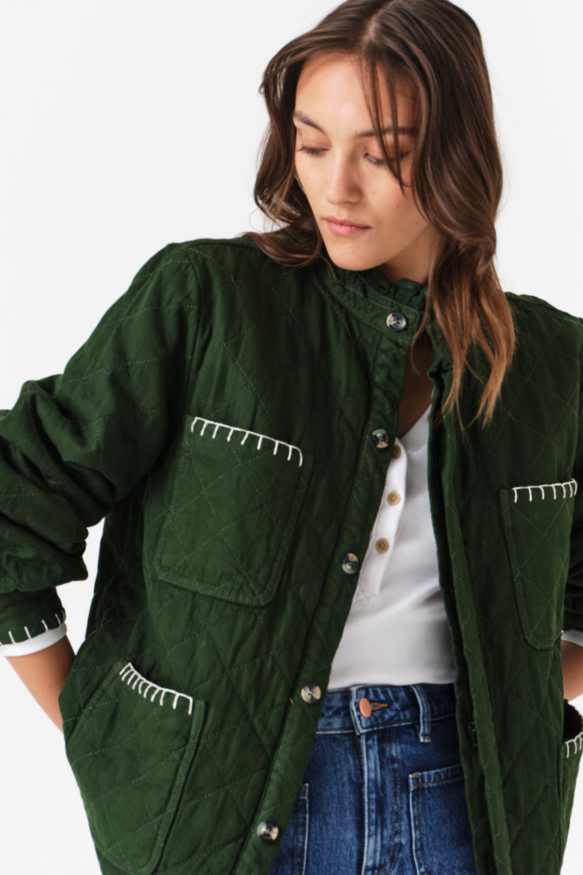 Close up of woman wearing a quilted dark green jacket with white embroidered details and blue jeans on a white background