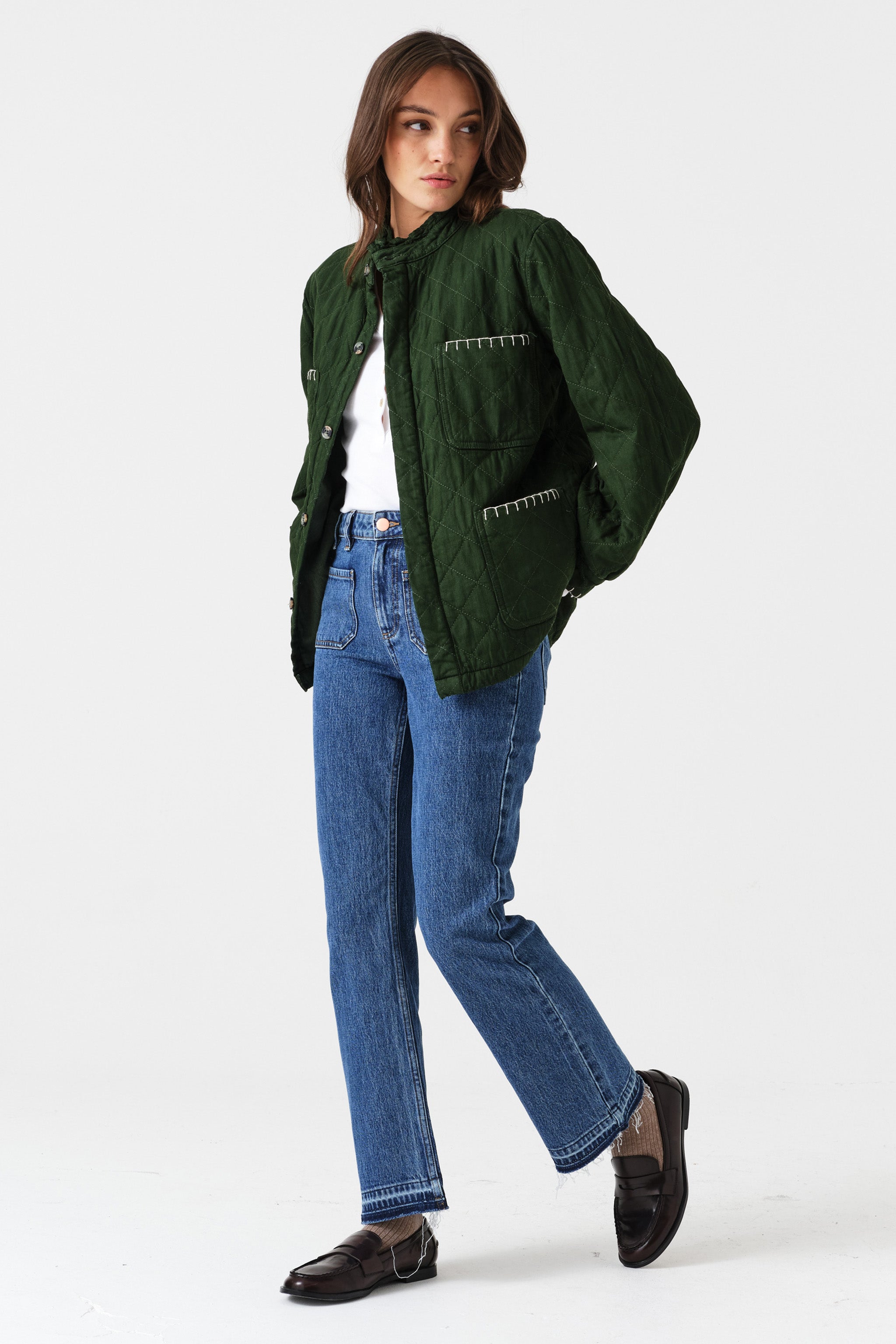 Woman wearing a quilted dark green jacket with white embroidered details and blue jeans on a white background